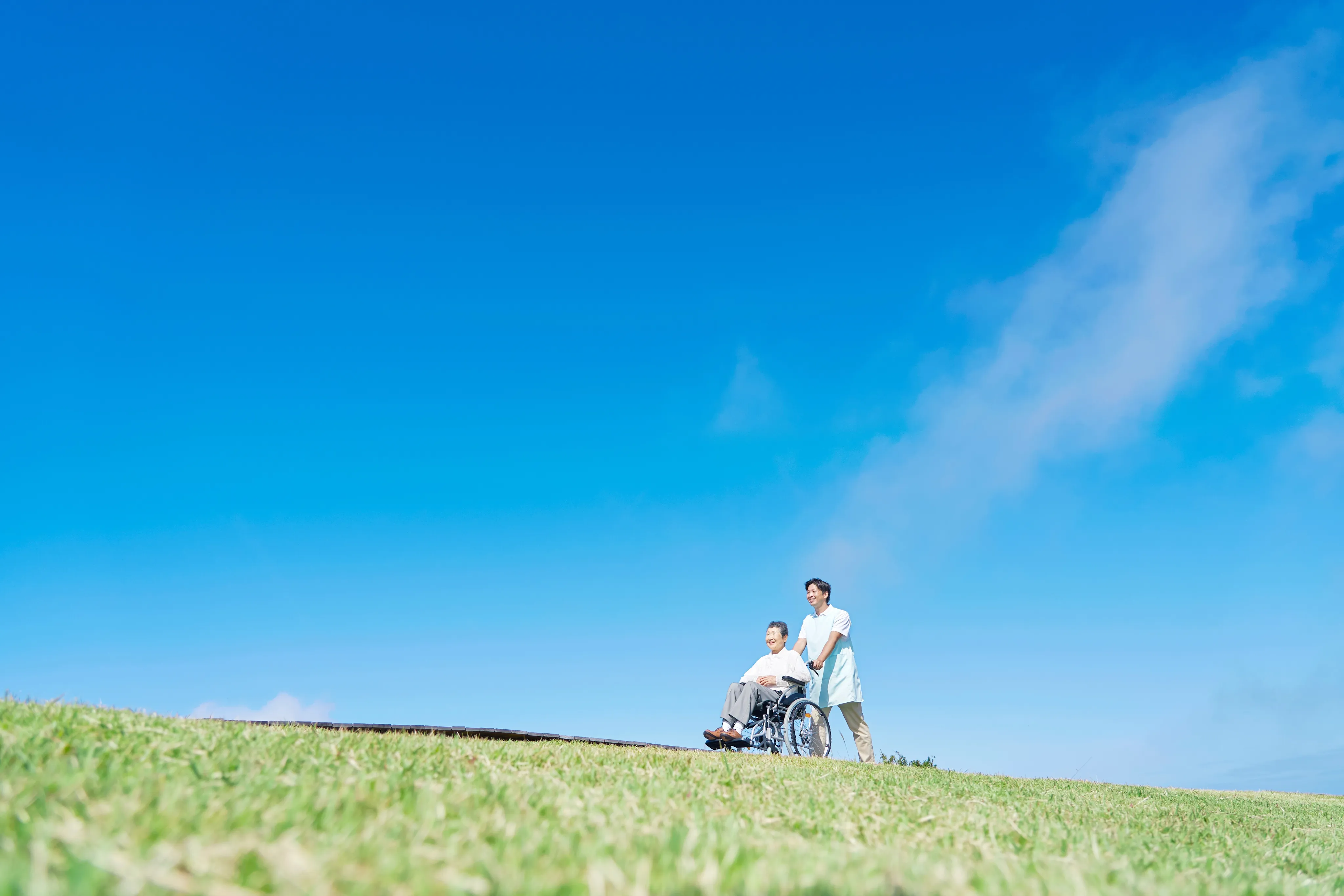 A caregiver pushing an older woman in a wheelchair across a grassy hill